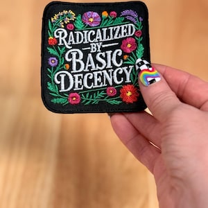 May include: A square black embroidered patch featuring the white text RADICALIZED BY BASIC DECENCY in the center. The text is surrounded by a colorful floral border with green leaves and various small flowers in shades of red, pink, purple, and orange.