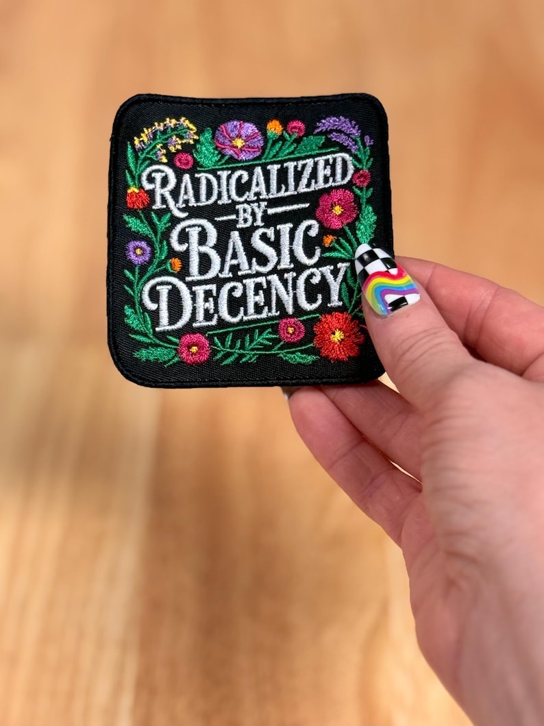 May include: A square black embroidered patch featuring the white text RADICALIZED BY BASIC DECENCY in the center. The text is surrounded by a colorful floral border with green leaves and various small flowers in shades of red, pink, purple, and orange.