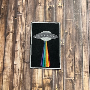 May include: An embroidered patch featuring a UFO with a rainbow beam of light emanating from its underside. The patch is set against a wooden backdrop.