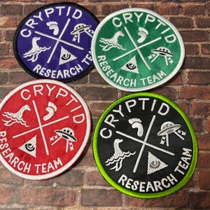 May include: Four embroidered patches with a white outline and the text "Cryptid Research Team". The patches are circular and feature a white footprint, a white UFO, a white eye, and a white sea monster on a purple, green, red, and black background.