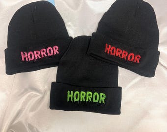 May include: Three black knit beanies with the word "HORROR" embroidered in different colors: pink, red, and green.
