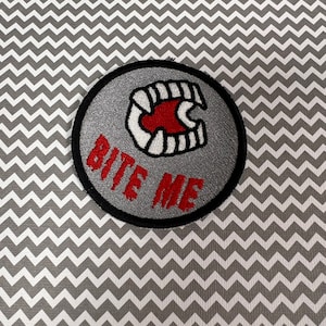 May include: A gray and red embroidered patch with a set of teeth and the words "Bite Me" in red.