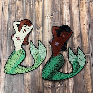 Op de afbeelding: Twee geborduurde strijkpatches van zeemeerminnen met groene staarten en stervormige bikini-tops. Eén zeemeermin heeft een bruine huid en de andere een witte huid.