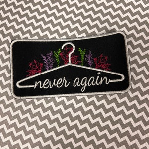 May include: Black embroidered patch with a white hanger and the text "never again" in white. The hanger is decorated with colorful flowers.