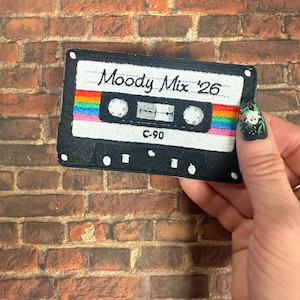 May include: A rectangular embroidered patch shaped like a cassette tape with a black frame and white center. The text Moody Mix '26 is stitched in black above a horizontal rainbow stripe. The center features two white circles and the text C-90.