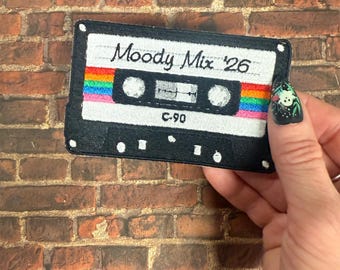 Puede incluir: Un parche bordado rectangular con forma de cinta de casete con un marco negro y un centro blanco. El texto Moody Mix '26 está bordado en negro sobre una franja horizontal de arcoíris. El centro presenta dos círculos blancos y el texto C-90.