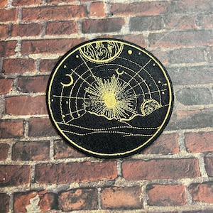 May include: A black circular patch with gold embroidery depicting a celestial scene. The design features a sun with rays, a crescent moon, and stars against a dark background.