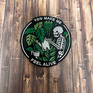 Könnte beinhalten: Schwarzer und weißer gestickter Patch mit einem Skelett, das Pflanzen gießt. Der Patch ist rund und hat den Text "You make me feel alive" am Rand.