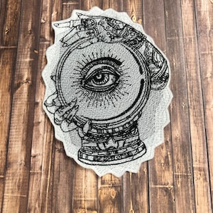 May include: A black and white embroidered patch of a hand holding a crystal ball with an eye inside. The hand is adorned with intricate details.
