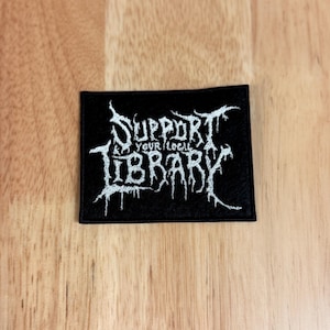 May include: Black rectangular patch with white embroidered text reading "SUPPORT YOUR LOCAL LIBRARY" in a gothic font. The patch is on a light brown wooden surface.