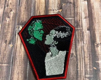 May include: Embroidered patch featuring a green and black Frankenstein's monster and a white and black Bride of Frankenstein in a red coffin shape.