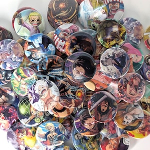 May include: A collection of round, colorful buttons featuring various anime characters. The buttons display a variety of vibrant illustrations, with a mix of character portraits and action scenes. The buttons are scattered across a white surface.