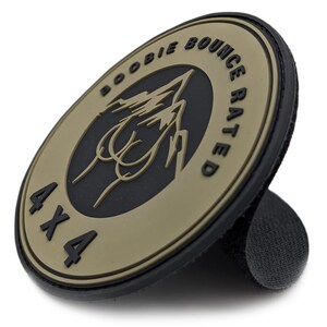 May include: A round, tan and black patch with the words "BOOBIE BOUNCE RATED" and "4X4" printed on it. The patch features a graphic of a mountain range and a stylized image. The patch has a hook and loop backing.