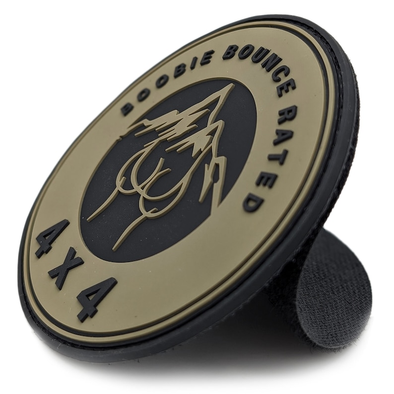 May include: A round, tan and black patch with the words "BOOBIE BOUNCE RATED" and "4X4" printed on it. The patch features a graphic of a mountain range and a stylized image. The patch has a hook and loop backing.