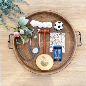 May include: A wooden tray with a black handle on each side. The tray contains a stethoscope, a gavel, a small white ball, a small notebook with a butterfly design, a blue cell phone, and a small wooden circle with coins on it.