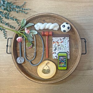 May include: A wooden tray with a variety of items, including a stethoscope, a gavel, a notebook with butterflies, a soccer ball, a ball of yarn, a small wooden log with coins, and a green smartphone. The tray has black metal handles.