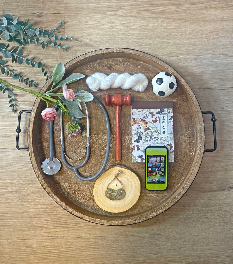May include: A wooden tray with a variety of items, including a stethoscope, a gavel, a notebook with butterflies, a soccer ball, a ball of yarn, a small wooden log with coins, and a green smartphone. The tray has black metal handles.