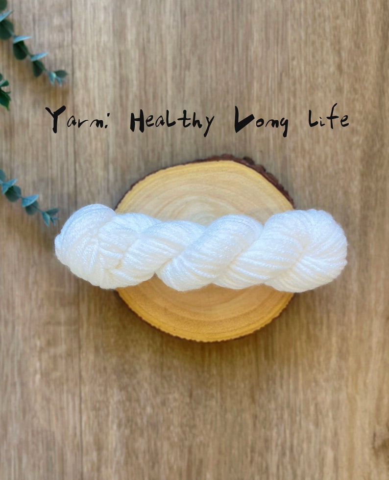 May include: White yarn on a wooden round surface. The yarn is twisted and has a slight sheen. The text "Yarn: Healthy Long Life" is written above the yarn.