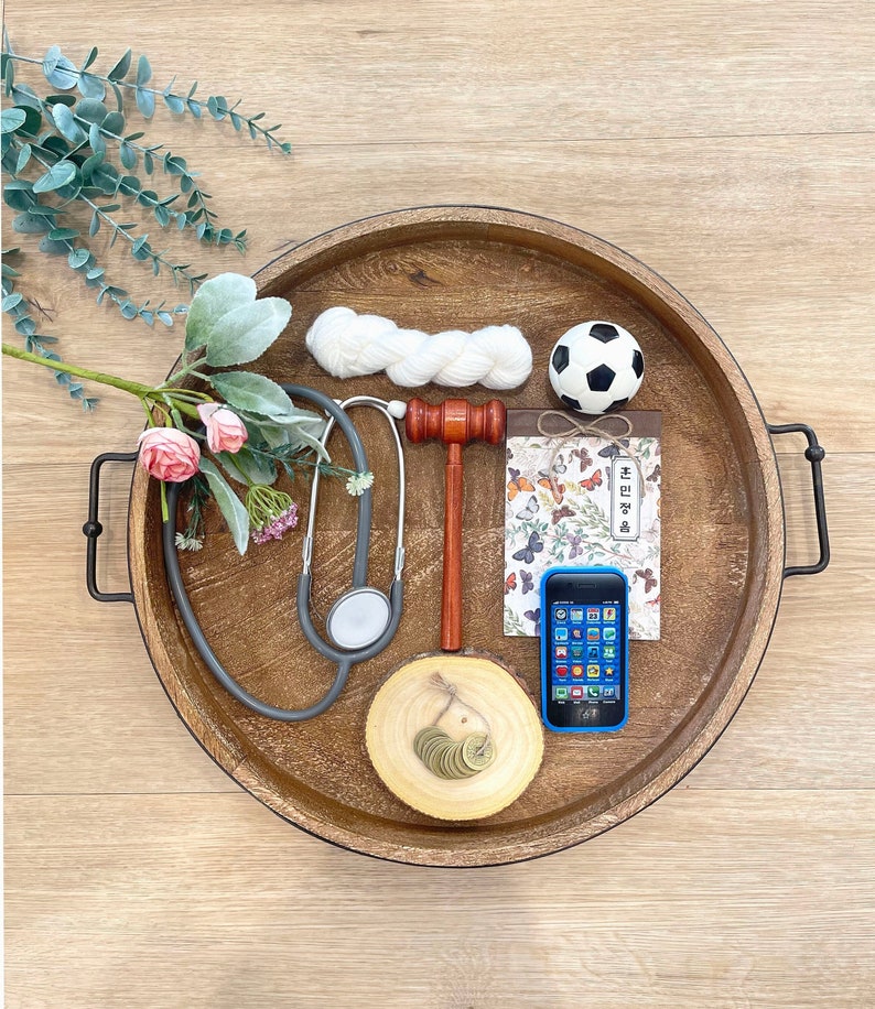 May include: A wooden tray with a black handle on each side. The tray contains a stethoscope, a gavel, a small white ball, a small notebook with a butterfly design, a blue cell phone, and a small wooden circle with coins on it.