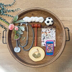 May include: A wooden tray with a variety of items, including a stethoscope, a gavel, a soccer ball, a notebook, a phone, and a white yarn ball. The tray is decorated with a floral design.