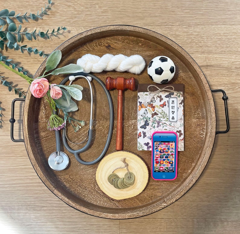 May include: A wooden tray with a variety of items, including a stethoscope, a gavel, a soccer ball, a notebook, a phone, and a white yarn ball. The tray is decorated with a floral design.