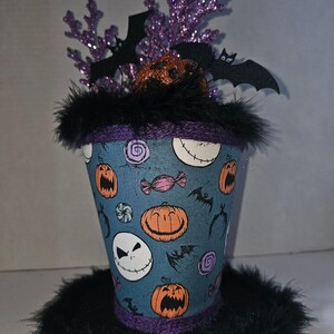 May include: A decorative top hat featuring a blue fabric covering with a pattern of orange jack-o'-lanterns, white skull faces, and colorful candies. The hat is trimmed with black feathers and topped with purple glittery branches and black bat cutouts.