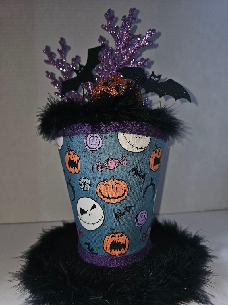 May include: A decorative top hat featuring a blue fabric covering with a pattern of orange jack-o'-lanterns, white skull faces, and colorful candies. The hat is trimmed with black feathers and topped with purple glittery branches and black bat cutouts.