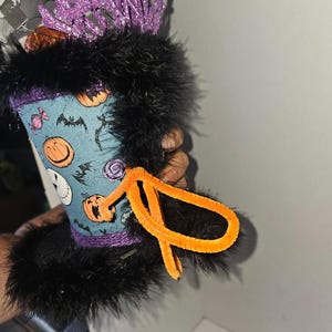 May include: A decorative top hat featuring a light blue fabric with a pattern of orange pumpkins, black bats, and candies. The hat is trimmed with black faux fur at the top and bottom, accented with a bright orange pipe cleaner bow and purple glittery branches.
