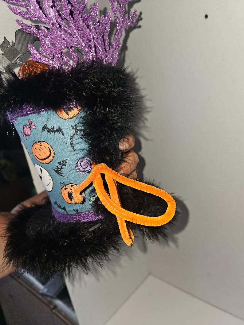 May include: A decorative top hat featuring a light blue fabric with a pattern of orange pumpkins, black bats, and candies. The hat is trimmed with black faux fur at the top and bottom, accented with a bright orange pipe cleaner bow and purple glittery branches.