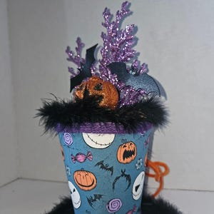 May include: A top hat decoration featuring a blue fabric base with a pattern of orange pumpkins, white skull faces, and colorful candies. The hat is trimmed with black feathers and topped with purple glittery branches, black bat cutouts, and a glittery orange pumpkin.