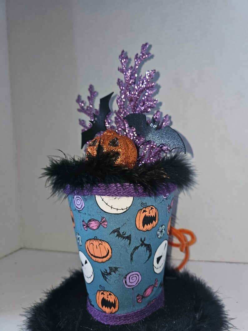 May include: A top hat decoration featuring a blue fabric base with a pattern of orange pumpkins, white skull faces, and colorful candies. The hat is trimmed with black feathers and topped with purple glittery branches, black bat cutouts, and a glittery orange pumpkin.
