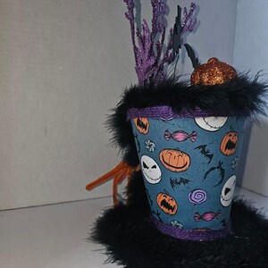 May include: A decorative top hat featuring a blue fabric base with a pattern of orange pumpkins, white skull faces, purple lollipops, and black bats. The hat is trimmed with black faux fur, purple rope, and topped with purple glittery sprigs.