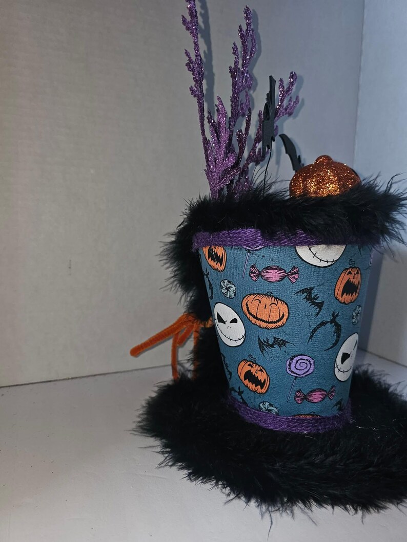 May include: A decorative top hat featuring a blue fabric base with a pattern of orange pumpkins, white skull faces, purple lollipops, and black bats. The hat is trimmed with black faux fur, purple rope, and topped with purple glittery sprigs.