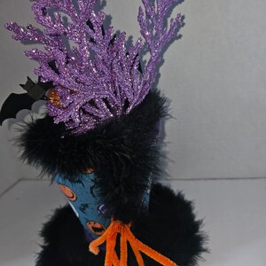 May include: A decorative cup featuring a blue exterior with orange pumpkin graphics, adorned with black faux fur trim at the top and base. The cup includes purple glittery branch-like accents, a black bat cutout, and orange pipe cleaners.