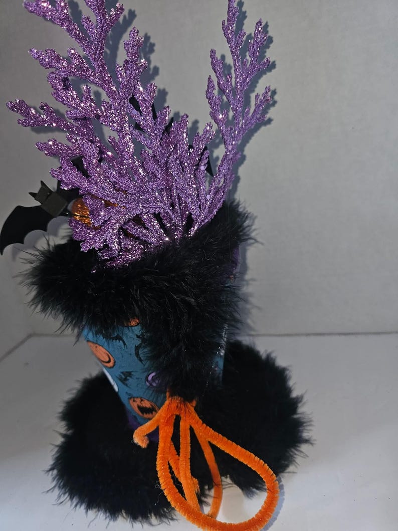May include: A decorative cup featuring a blue exterior with orange pumpkin graphics, adorned with black faux fur trim at the top and base. The cup includes purple glittery branch-like accents, a black bat cutout, and orange pipe cleaners.