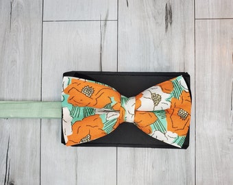 May include: A bow tie with a floral pattern featuring orange and white flowers on a teal background. The bow tie is on a light wood surface.