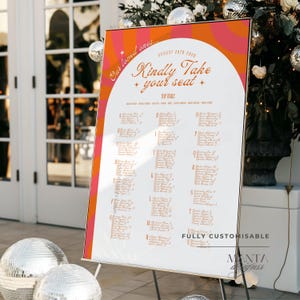May include: A wedding seating chart sign featuring a white central arch with orange and pink radial stripes. The text reads "Our loved ones...", "Kindly Take your seat", "AUGUST 28TH 2028", and "TOP TABLE" with a list of names organized by letter.