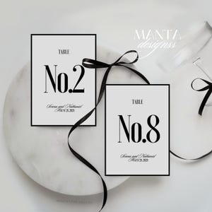 以下が含まれることがあります： 明るい灰色の背景に、黒い数字「No.2」と「No.8」が描かれた2枚のエレガントなテーブルナンバーカード。各カードは黒いフレームで囲まれ、「Serena and Nathaniel」の名前と「August 28, 2028」の日付が含まれています。黒いリボンが装飾的なアクセントになっています。