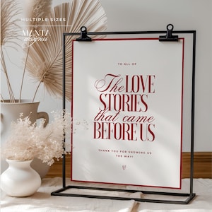 May include: A framed print with the text "The LOVE STORIES that came BEFORE US" in burgundy script. The print is held by a black metal stand. Decorative vases and dried plants are in the background.