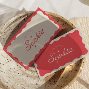 Puede incluir: Dos tarjetas de lugar rojas y blancas con el texto "Mesa Dos Sophia" en una fuente cursiva. Las tarjetas están sobre un plato marrón moteado.