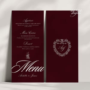 May include: Burgundy wedding menu with white script text. The menu lists an appetizer of roasted butternut squash soup, a main course of mushroom risotto, and a dessert of classic vanilla panna cotta. The cover features a heart-shaped design with initials.