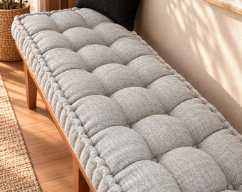 May include: A long wooden bench featuring a thick, light gray tufted cushion with a textured fabric finish. The cushion is secured with button detailing and has a piped edge. A single black square throw pillow rests on the bench seat.