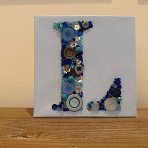 May include: A light blue canvas features the letter "L" crafted with various blue buttons, beads, and sequins. The decorative artwork is mounted on a wooden surface, showcasing a handmade, personalized design.