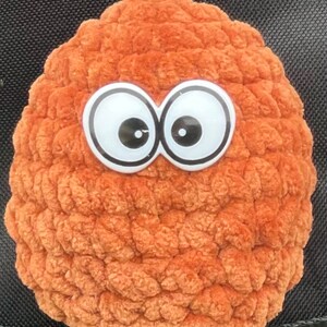 Pode incluir: Um brinquedo de pelúcia marrom crochetado com grandes olhos saltados brancos. O brinquedo tem a forma de uma bola e uma superfície texturizada.
