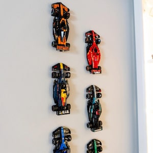 May include: A collection of colorful toy race cars mounted on a wall. The cars are various colors, including black, orange, red, blue, and green. Each car has detailed features and sponsor logos. The cars are arranged in a vertical pattern.