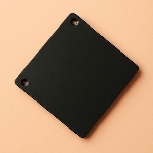 Op de afbeelding: Een zwart vierkant object met afgeronde hoeken en twee kleine gaten. Het object heeft een matte afwerking en staat tegen een perzikkleurige achtergrond. Het object is effen van kleur en lijkt van metaal of een vergelijkbaar materiaal te zijn.