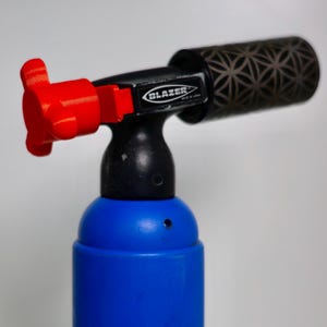 May include: A blue and black Blazer torch with a red control knob. The torch has a patterned design on the end and the brand name is visible. Made in Japan is printed on the side.