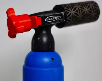 May include: A blue and black Blazer torch with a red control knob. The torch has a patterned design on the end and the brand name is visible. Made in Japan is printed on the side.