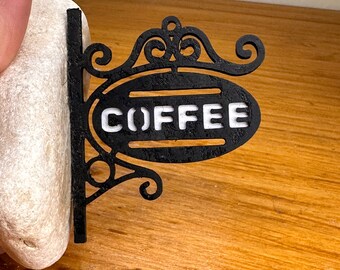 May include: A black metal sign with the word "COFFEE" in white, set against a wooden background. The sign has an ornate, decorative design with scrollwork and an oval shape. The sign is designed to hang.