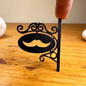 Puede incluir: Un letrero de metal negro con un diseño decorativo de volutas y un recorte de un bigote. El letrero está unido a un poste de metal negro.